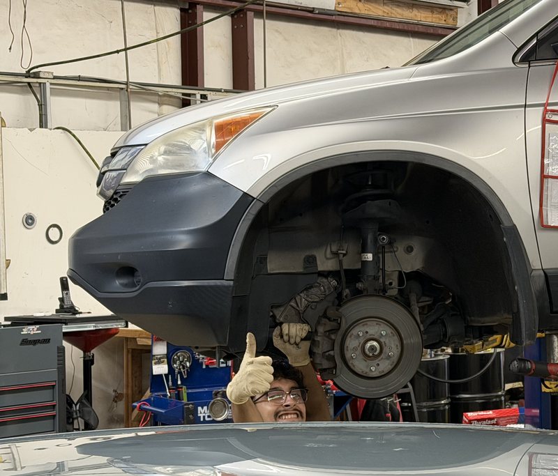 A smiling technician gives a thumbs up while working on the front brakes of an SUV on a lift