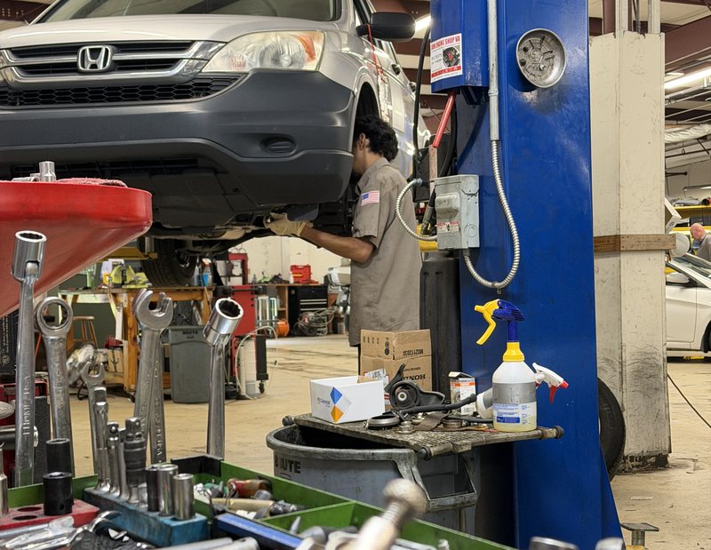 A technician reaches under a Honda SUV raised on a lift, with wrenches and sockets in the foreground