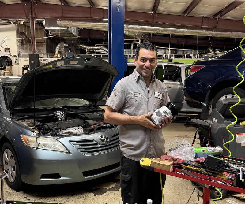Technician Murad smiles while holding a new air-conditioning compressor next to a Toyota with its hood open